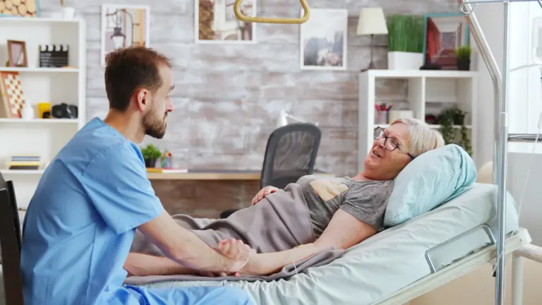A caregiver sitting beside an older woman and holding her hand.