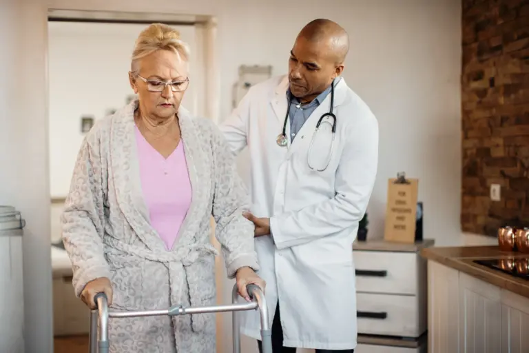A clinician supporting an older woman using a walking frame.