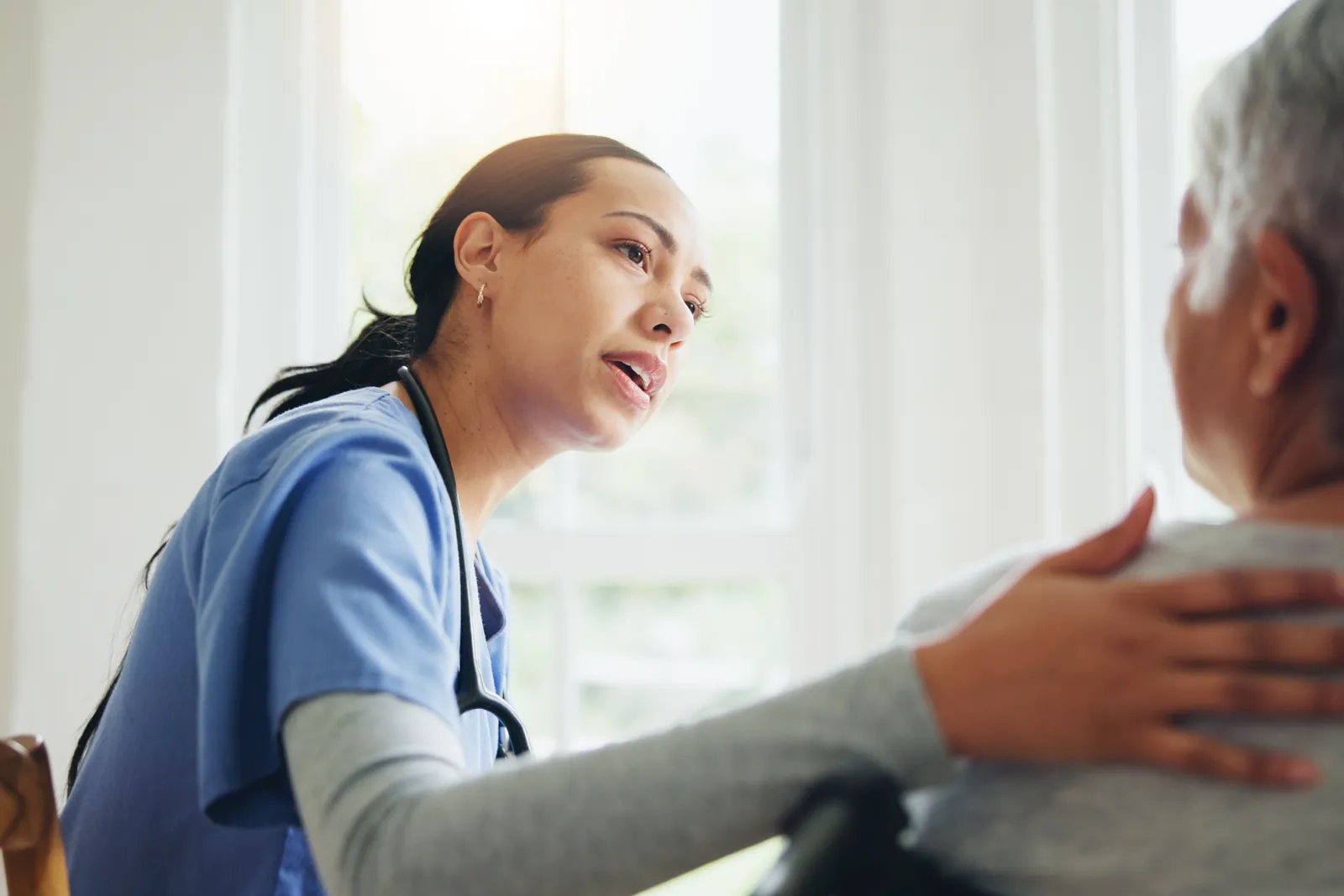 A nurse leans in close and speaks gently to an older person in a wheelchair, with a hand on their back.