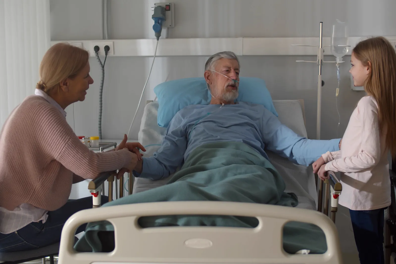 An older man in a hospital bed, on oxygen, holds hands with a visiting relative. A young girl stands by the bed.