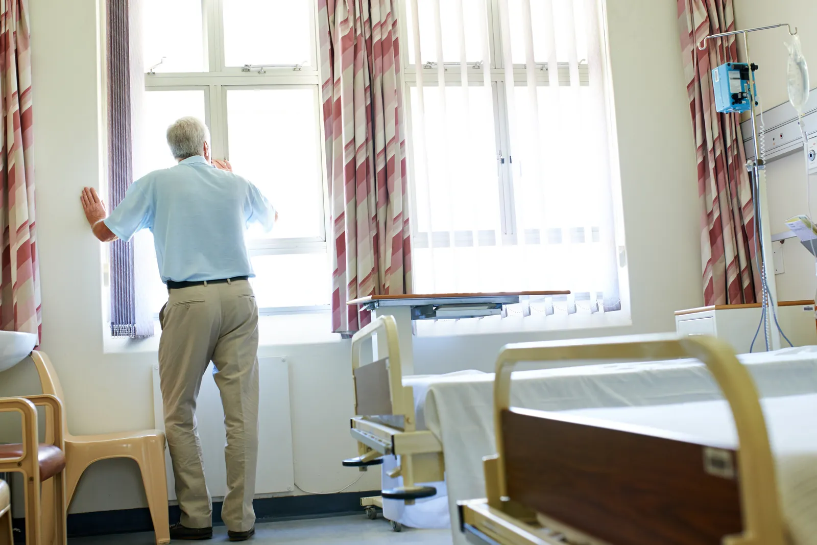 An older man stands at the window of a hospital room with his hand on the frame.