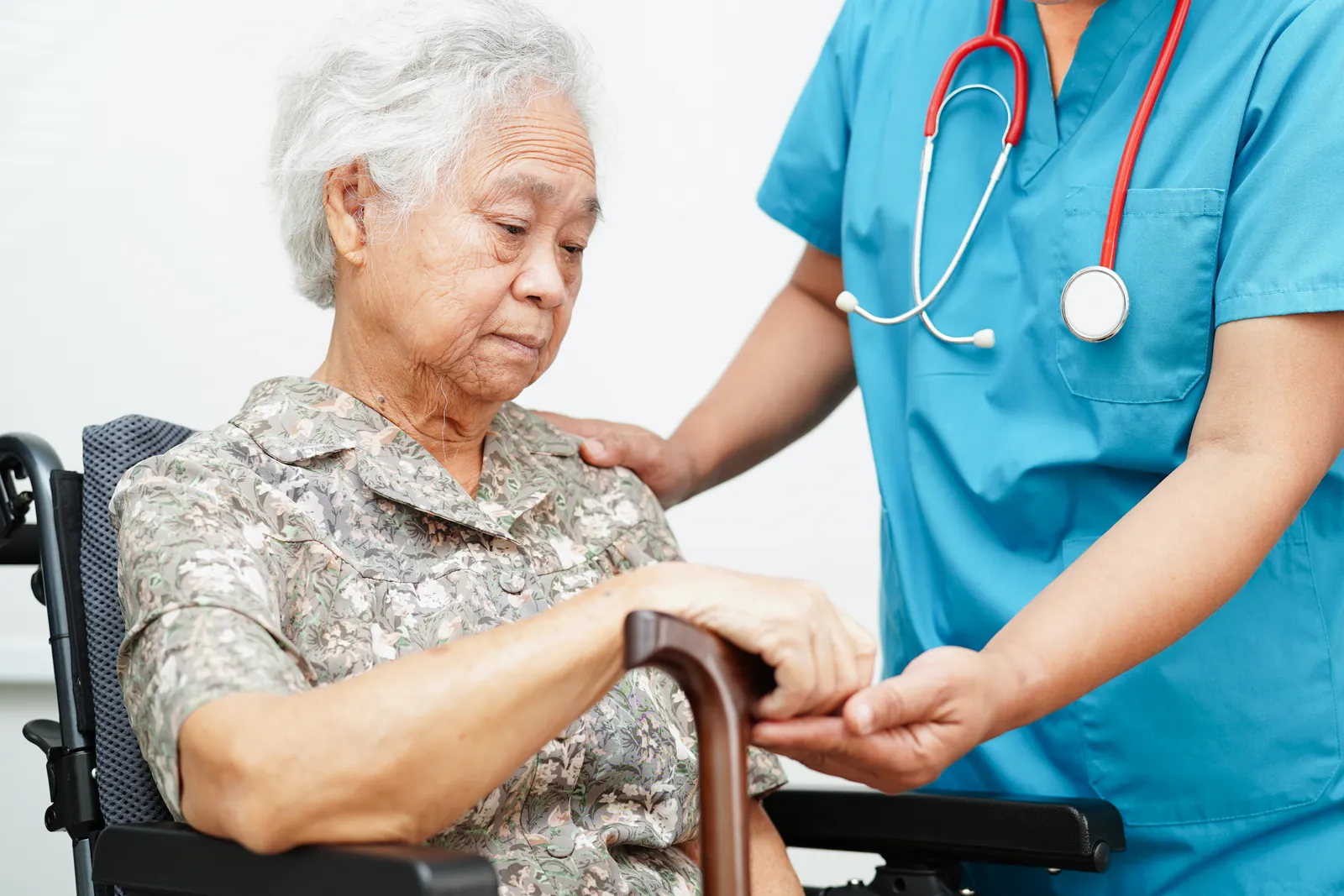 A nurse holds the hand of an older woman in a wheelchair, who looks down, worried.
