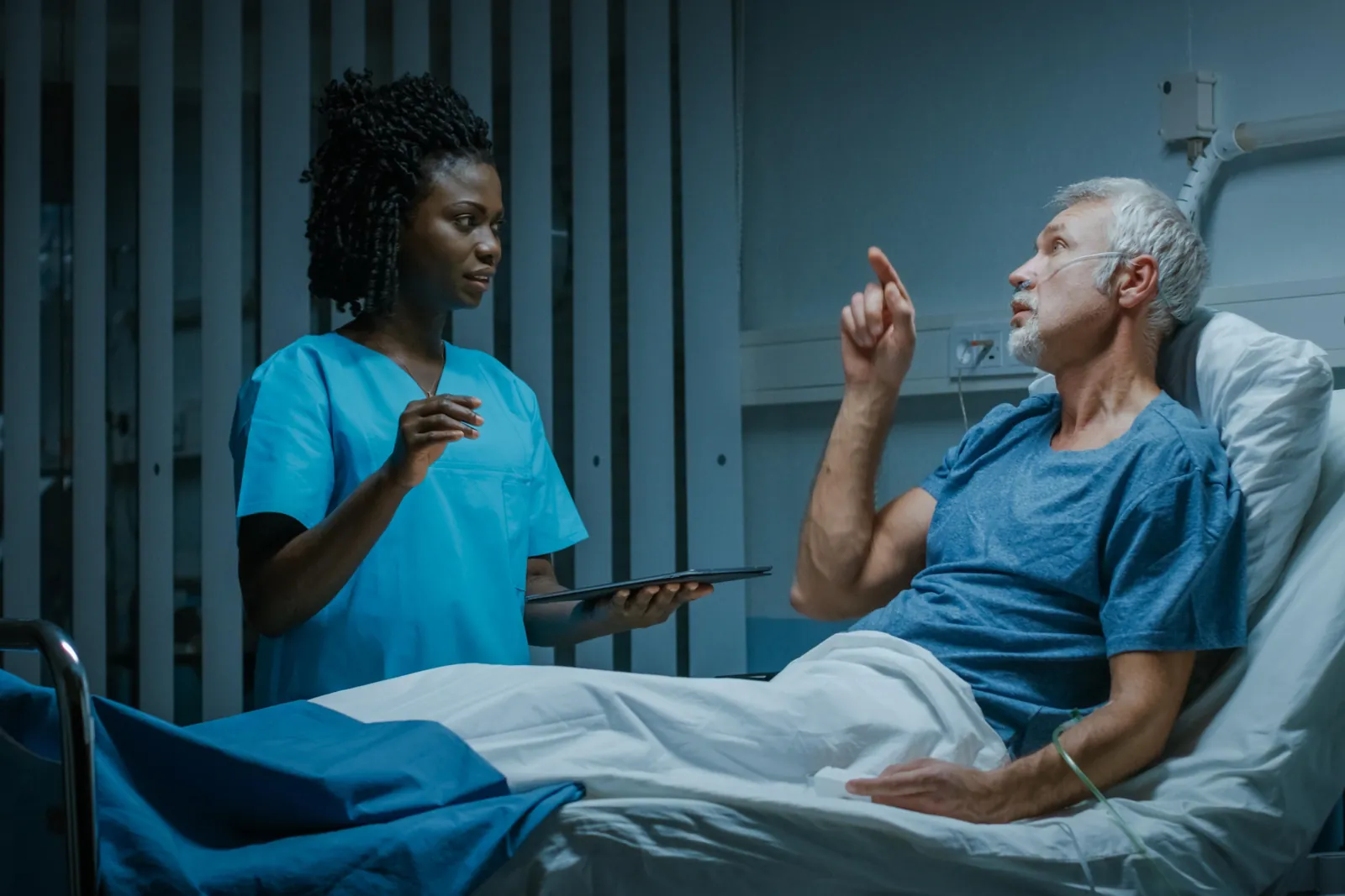 At night on a ward, an older man sits up in bed, pointing and talking. A nurse stands by the bed and listens.