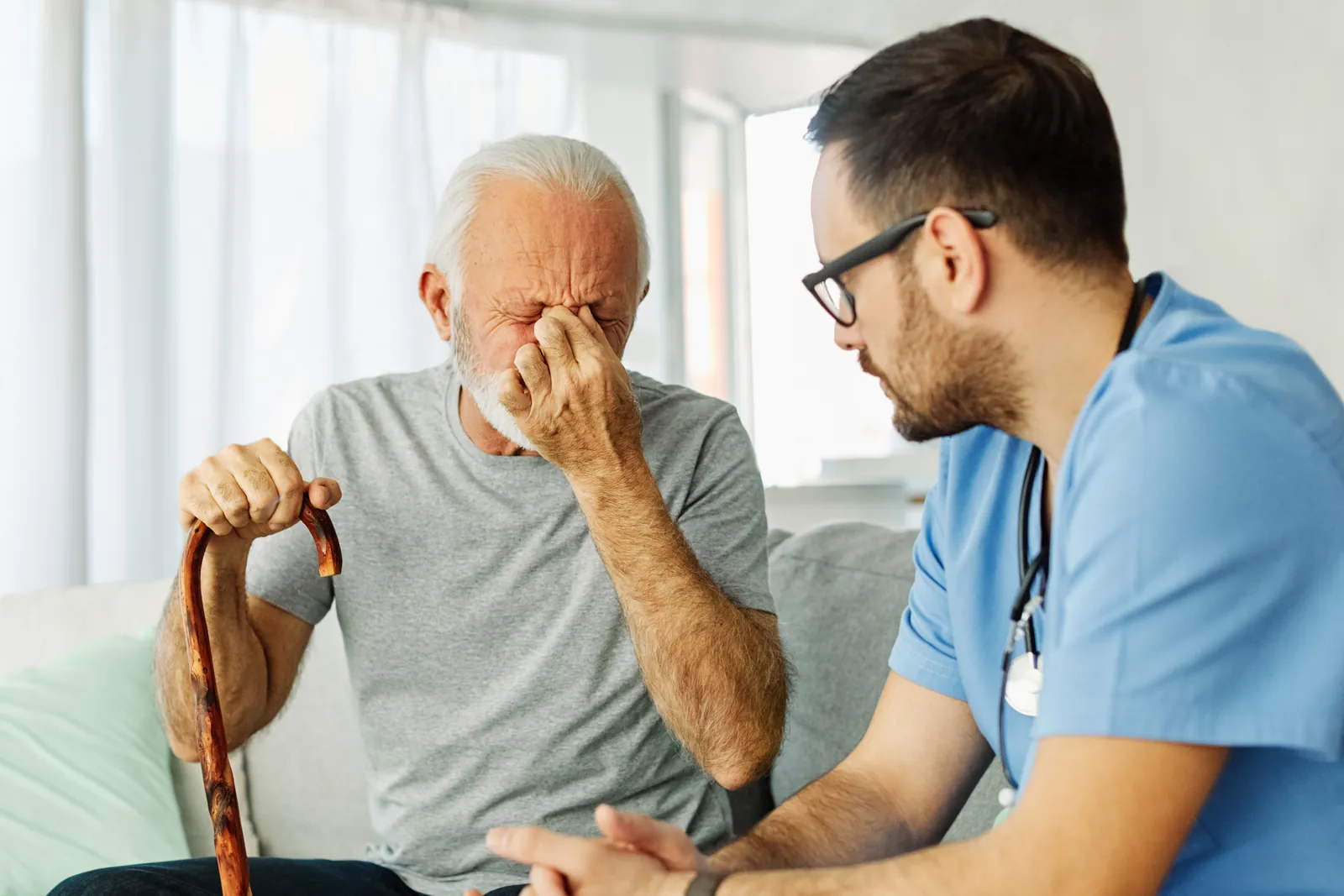 A male nurse leans in and listens to an older man who is upset, with his hand over his eyes.