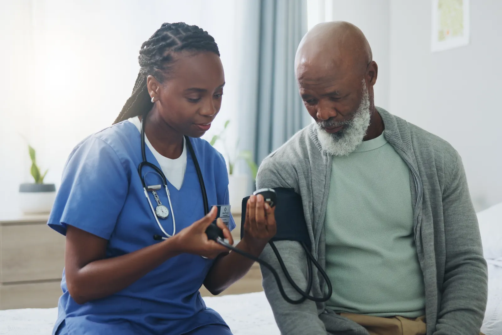 A nurse checks the blood pressure of an older man who is sitting on his bed.