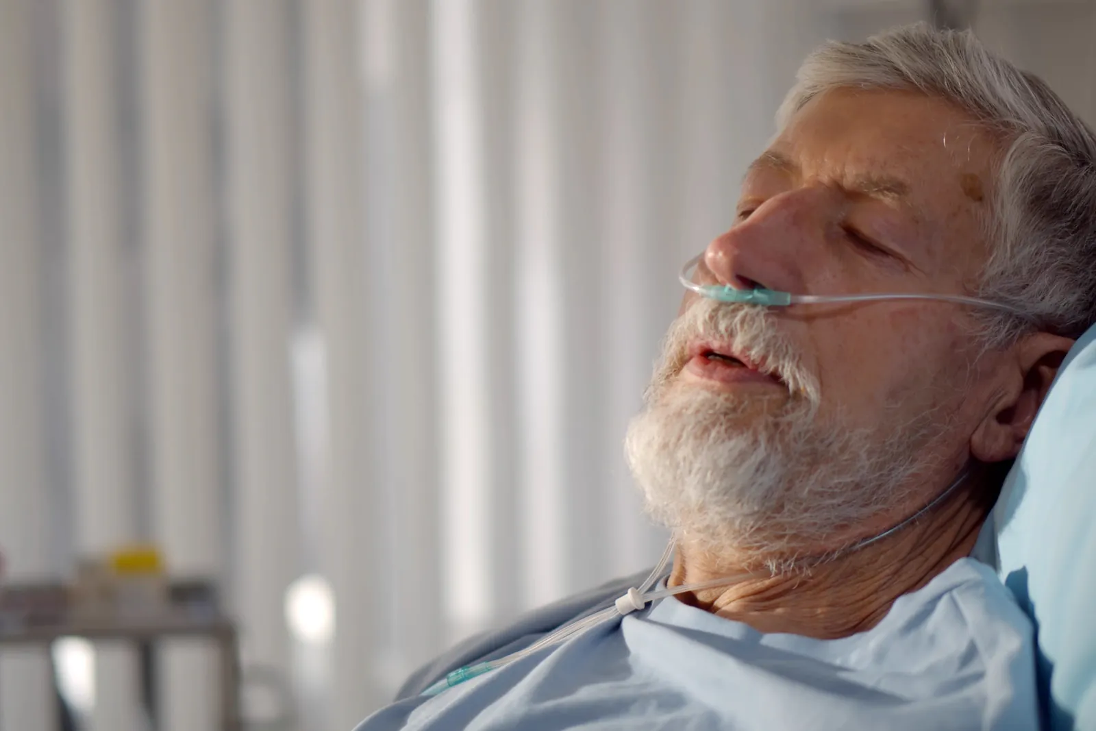 An older man lies in a hospital bed with oxygen tubing. His eyes are almost closed.