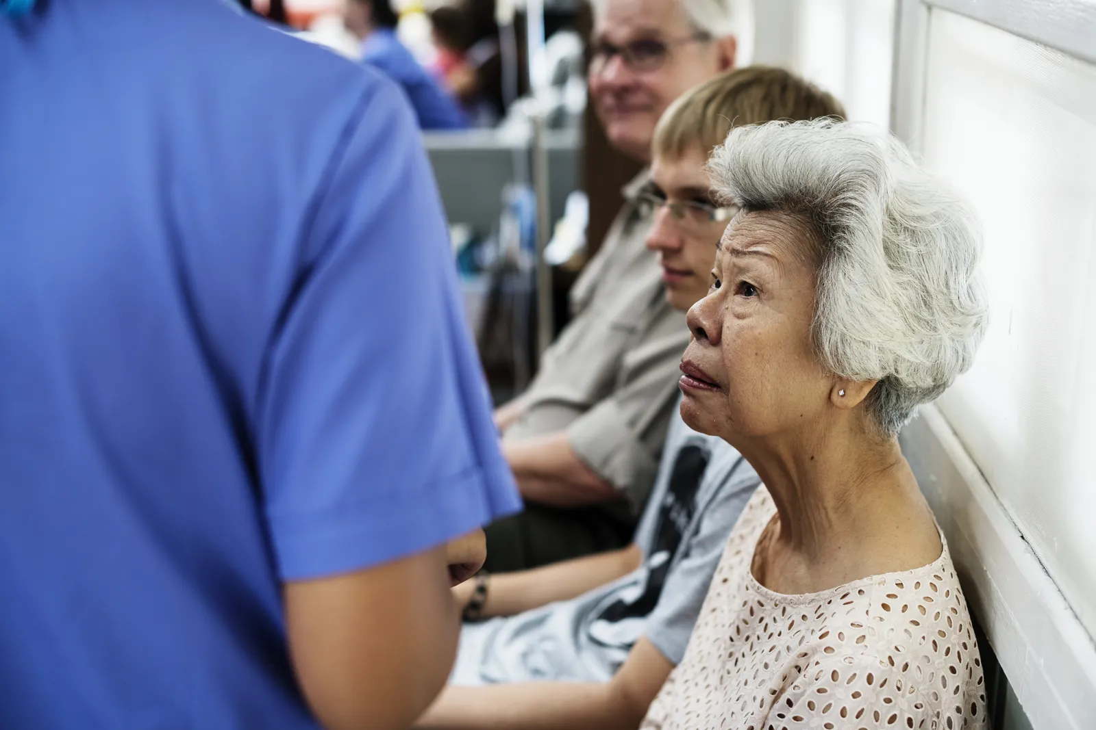 In a busy hospital bay, an older woman looks up at a nurse with a worried face.
