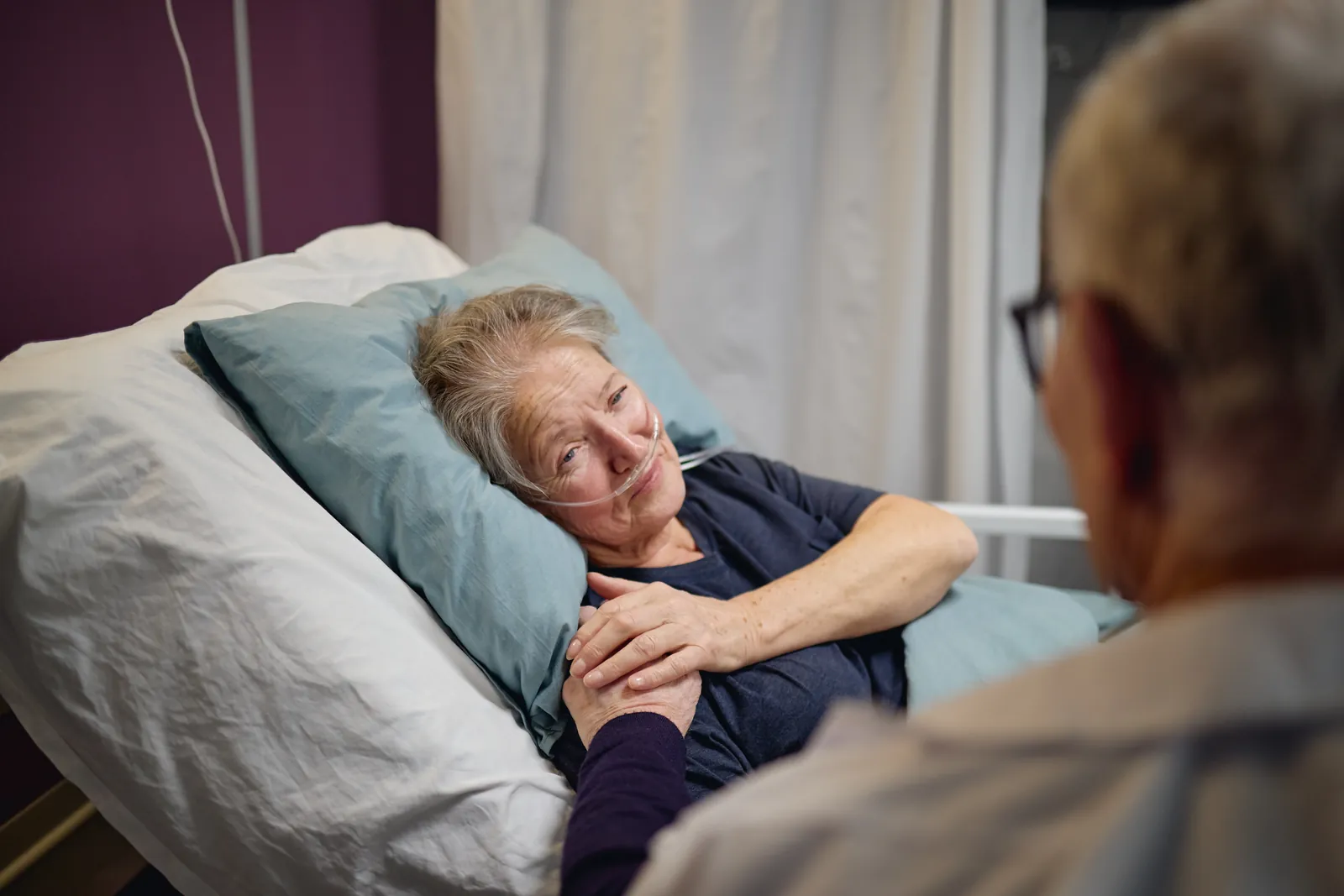 A drowsy older woman lies in a hospital bed with oxygen tubing. A visitor holds her hand.