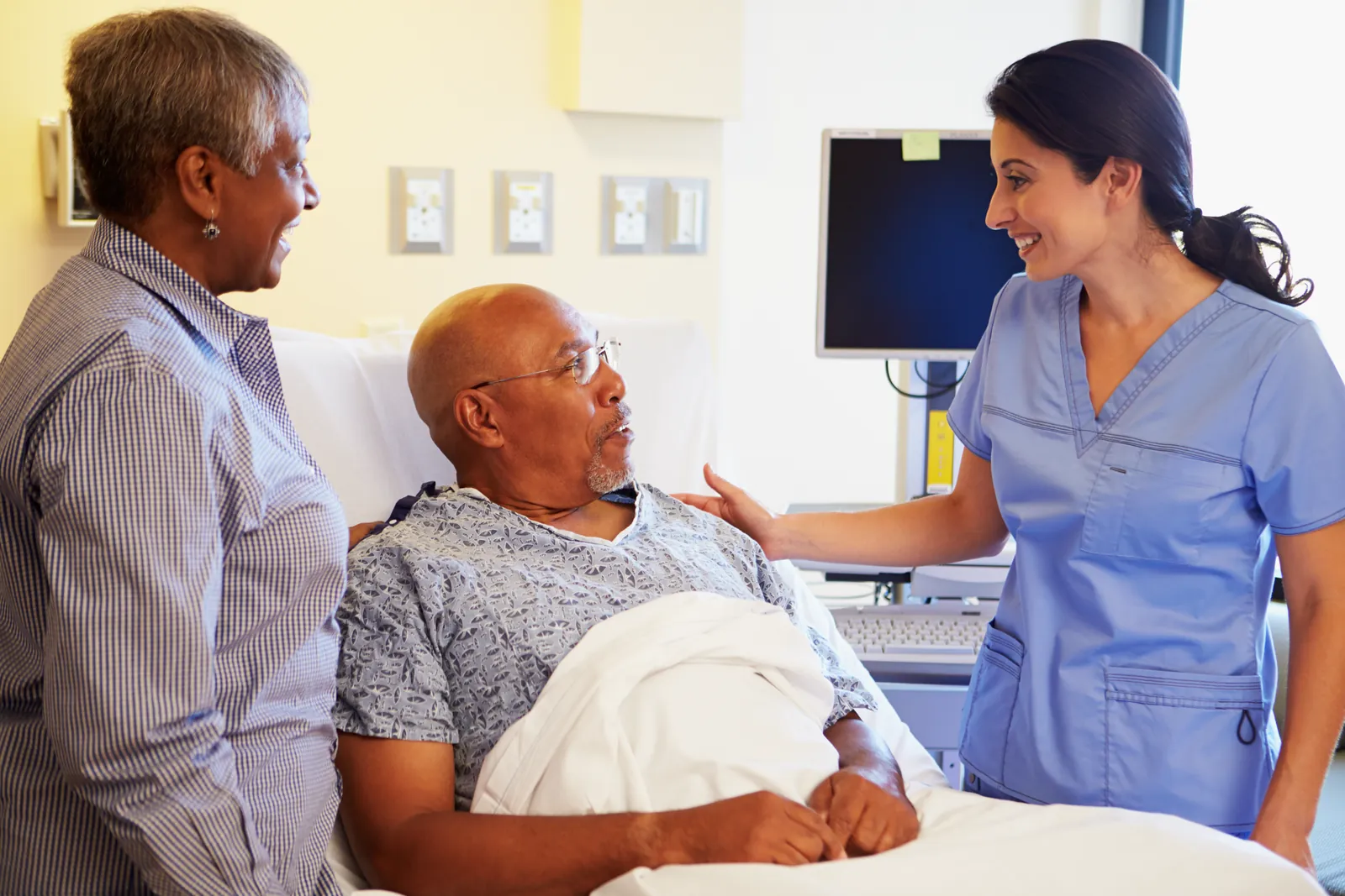 A nurse talks with an older man in a hospital bed and his wife, who stands beside him.