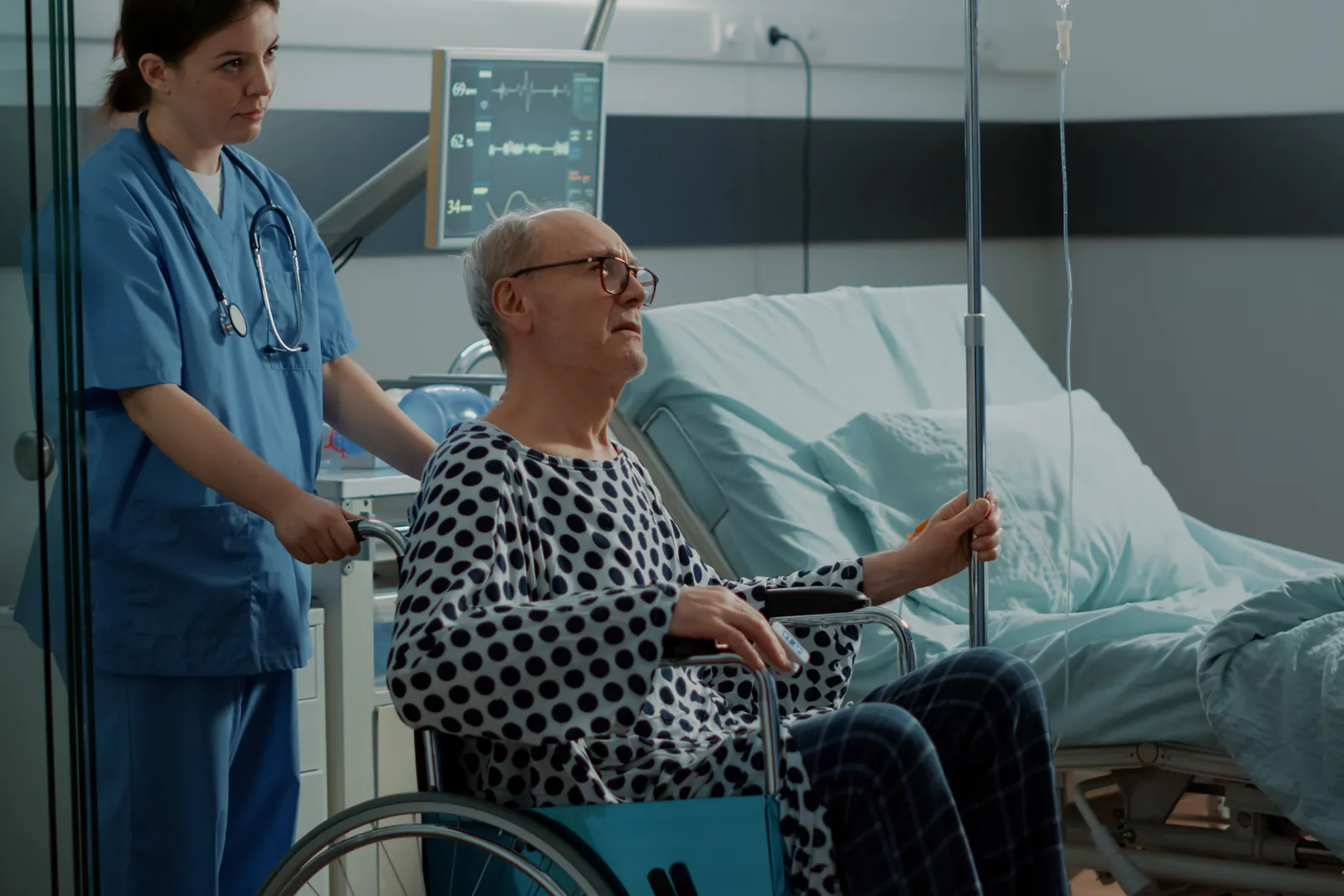 A nurse wheels an older man in a hospital gown out of the ward while a doctor checks his drip.