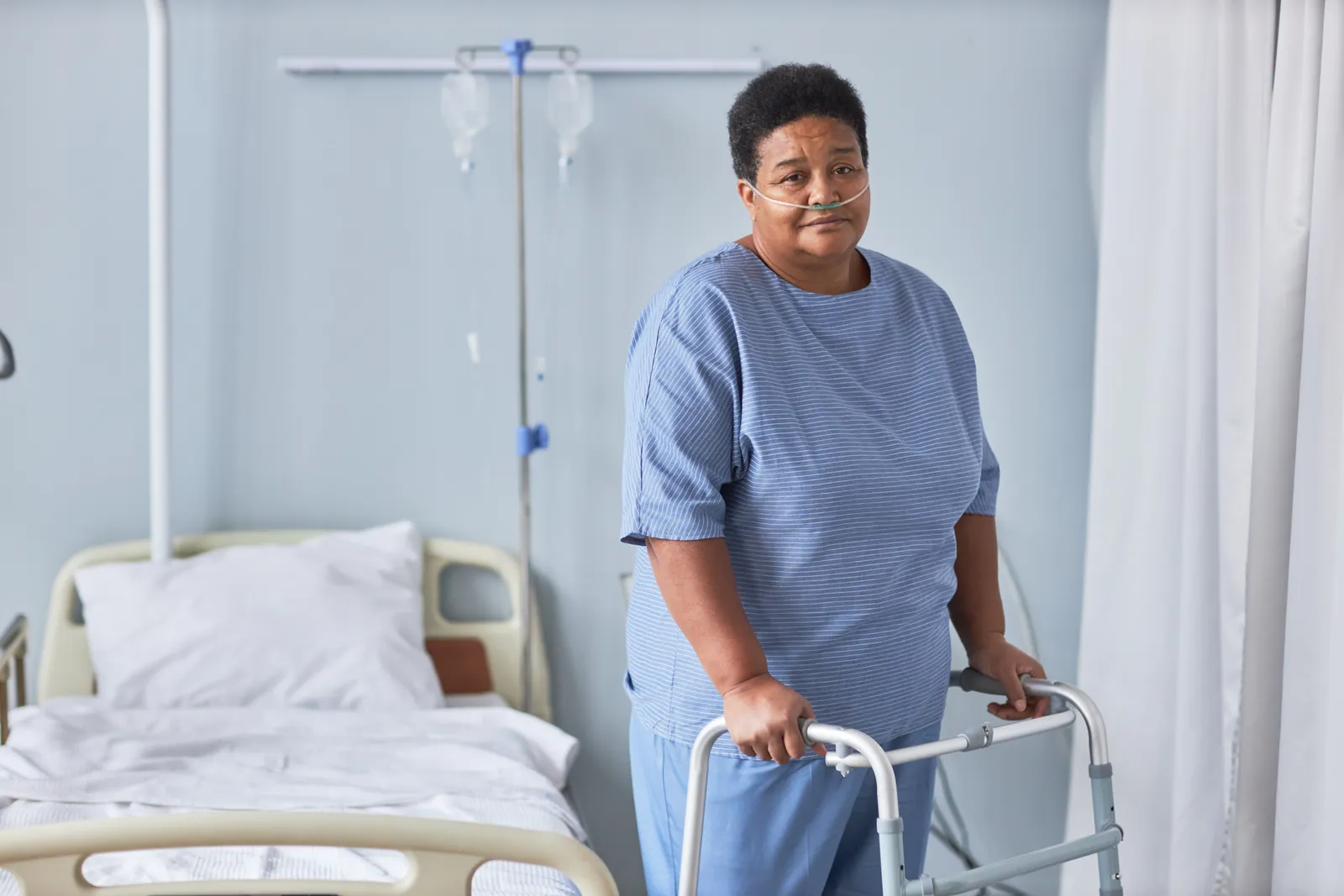 An older woman with oxygen tubing stands beside her hospital bed, holding a walking frame.