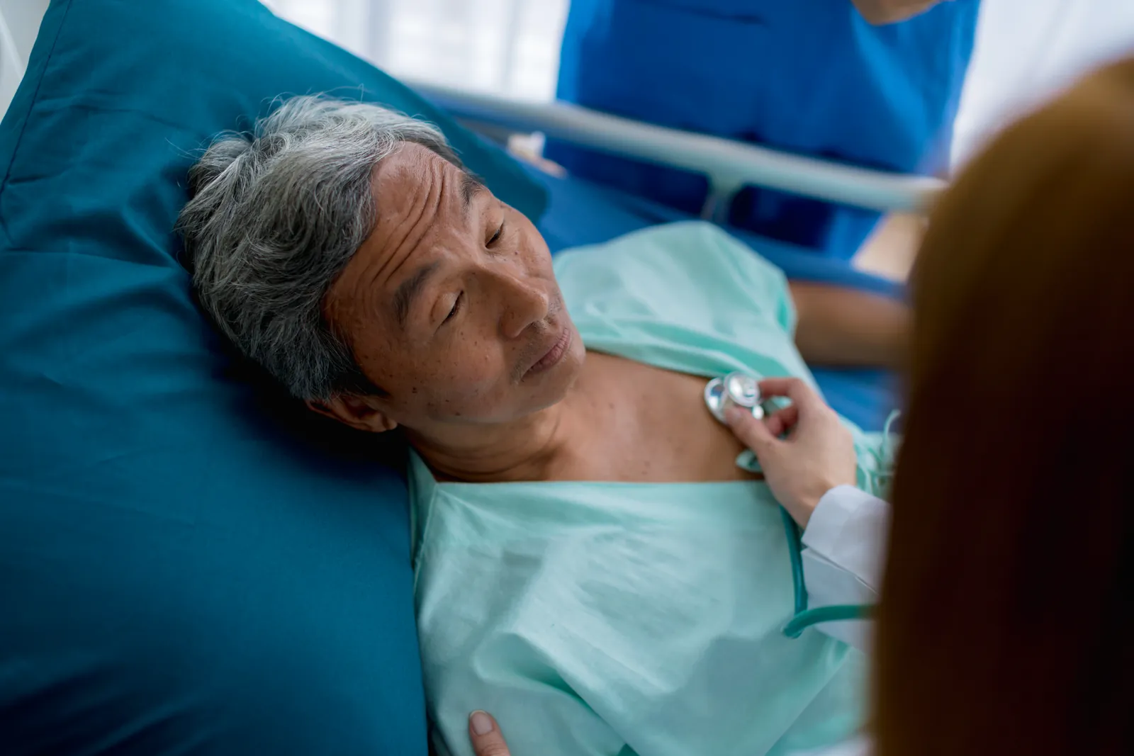 A clinician listens to the chest of an older man lying in a hospital bed.
