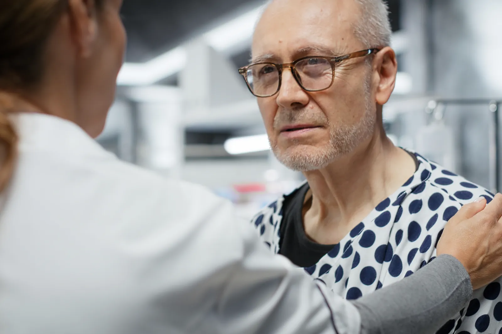 A clinician rests a hand on the shoulder of an older man in a spotted hospital gown.
