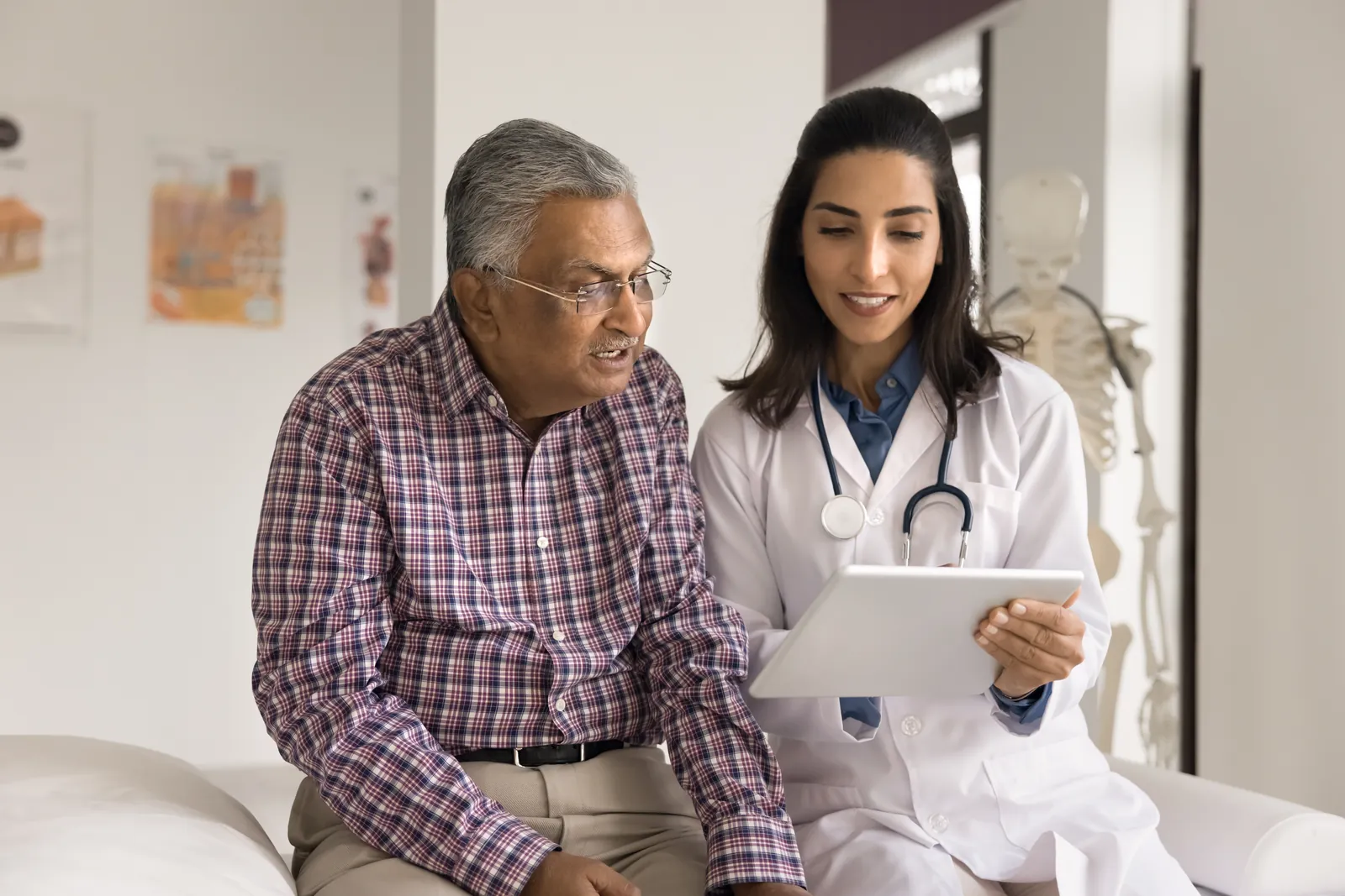 A doctor sits beside an older man and goes through information on a tablet with him.
