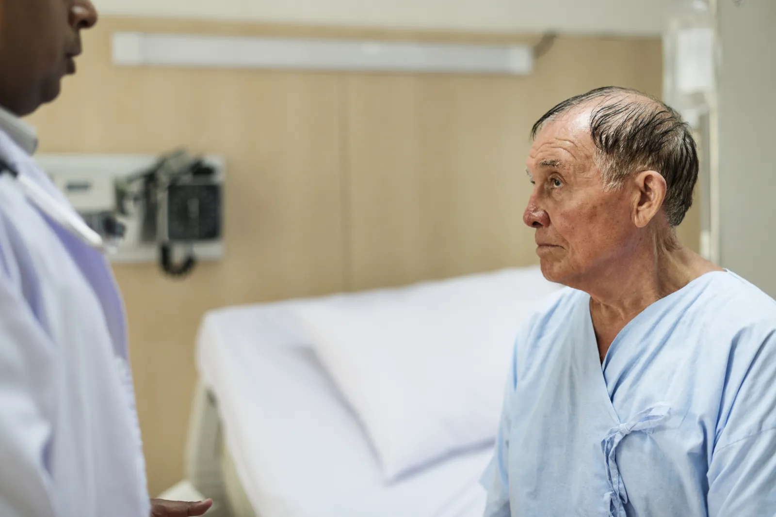 An older man in a hospital gown sits on his bed and looks uncertain while a clinician stands nearby.