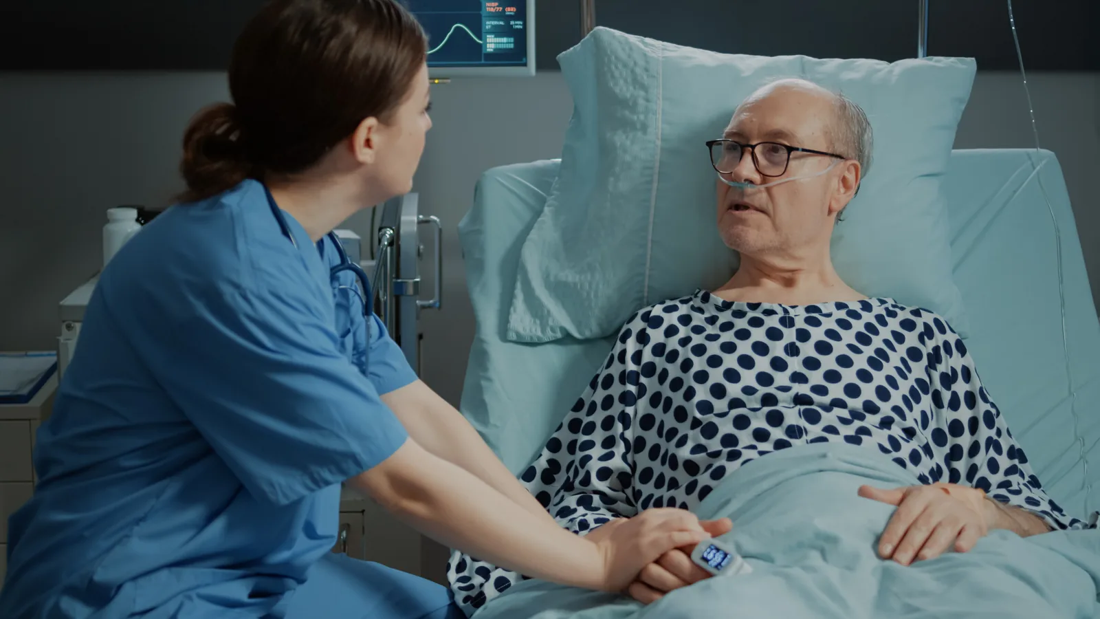 A nurse sits beside an older man in a hospital bed and holds his hand while he talks. He has oxygen tubing in his nose.