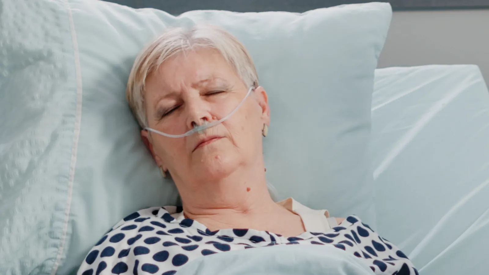 An older woman lying in a hospital bed with her eyes closed and nasal oxygen in place.