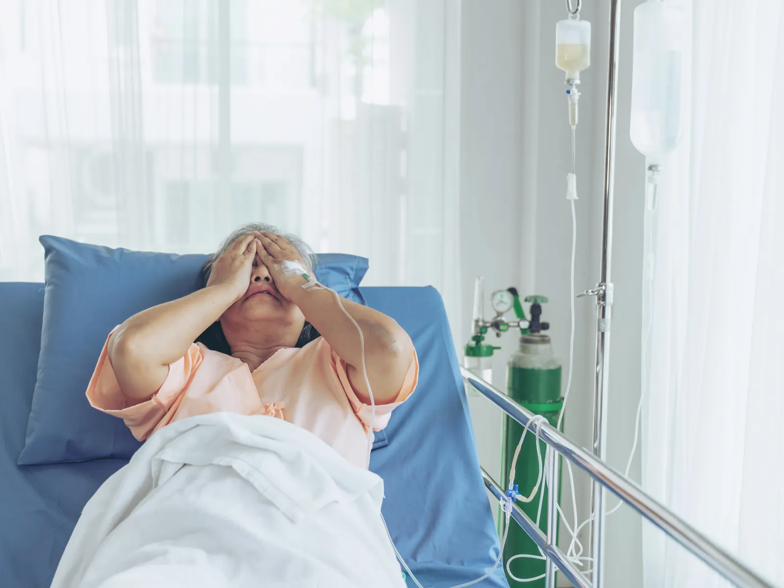 An older woman in a hospital bed with both hands over her face.