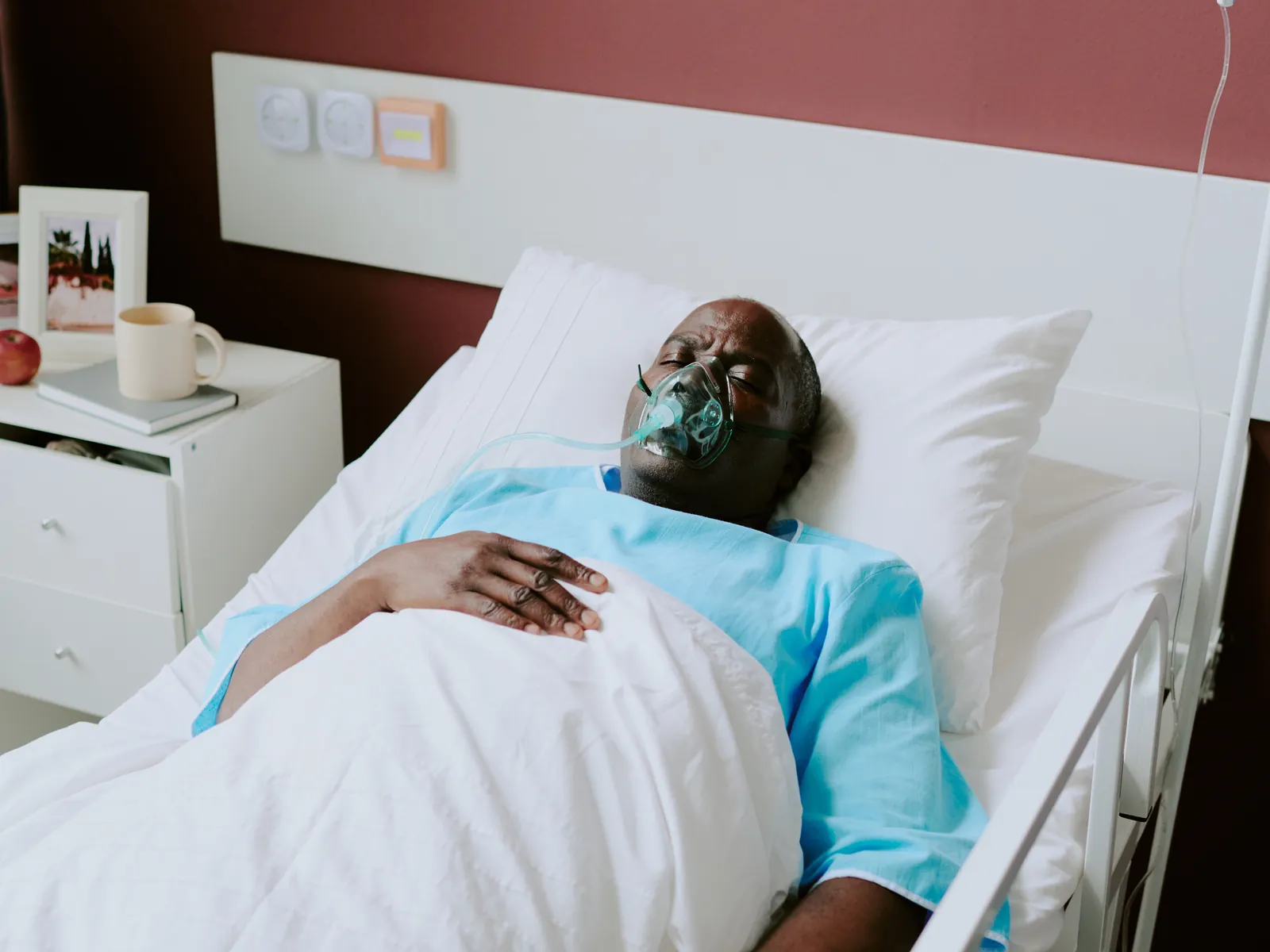 An older man resting in a hospital bed with his eyes closed and an oxygen mask in place.