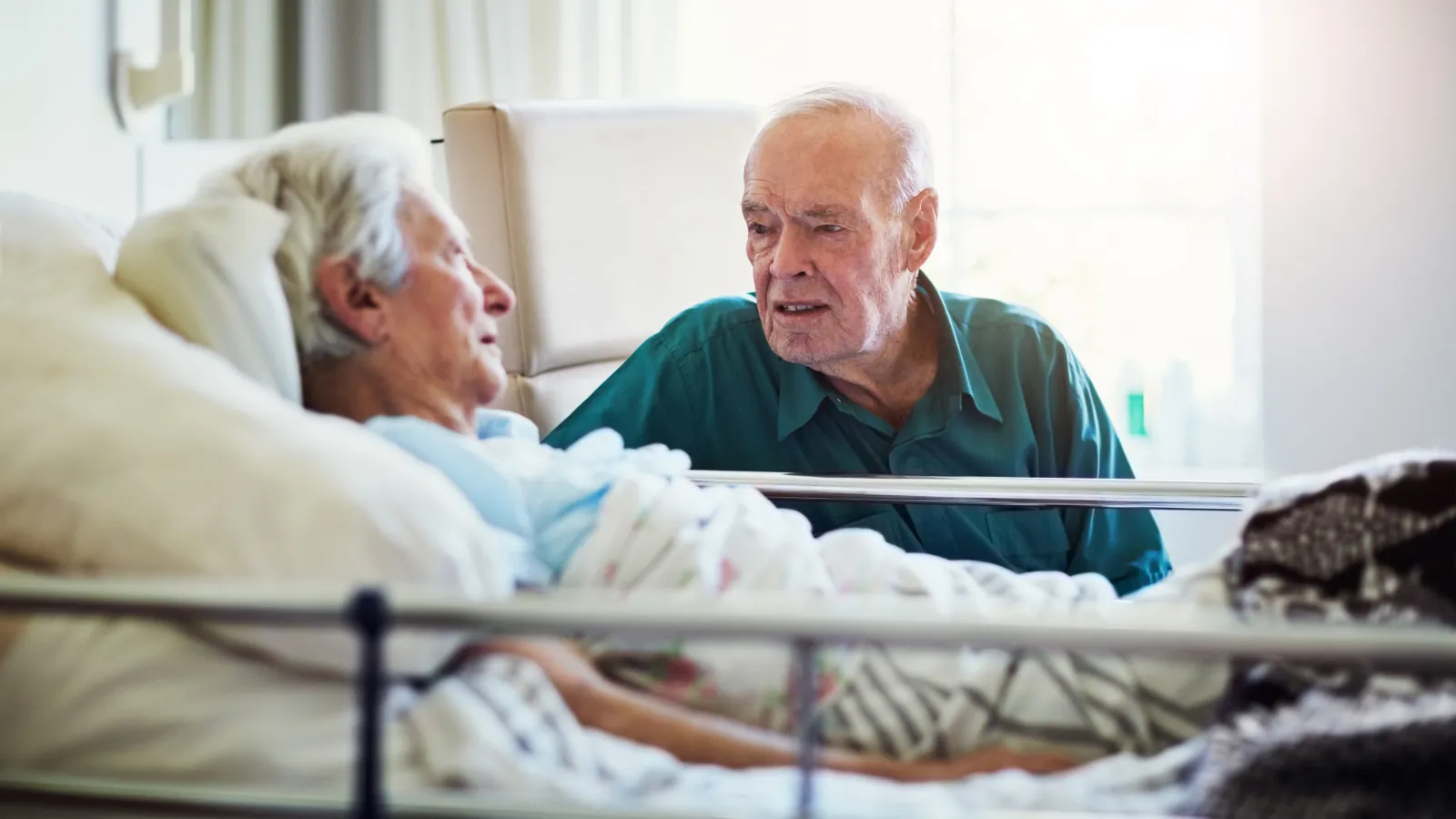 An older man sitting beside his wife's hospital bed, talking with her.