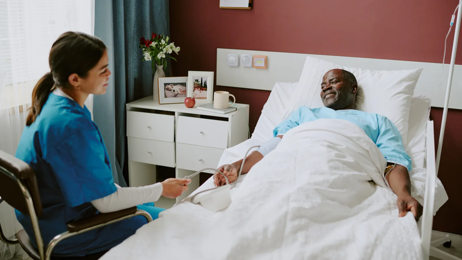 A nurse seated at eye level beside an older man in a hospital bed; a framed family photograph stands on the bedside cabinet.