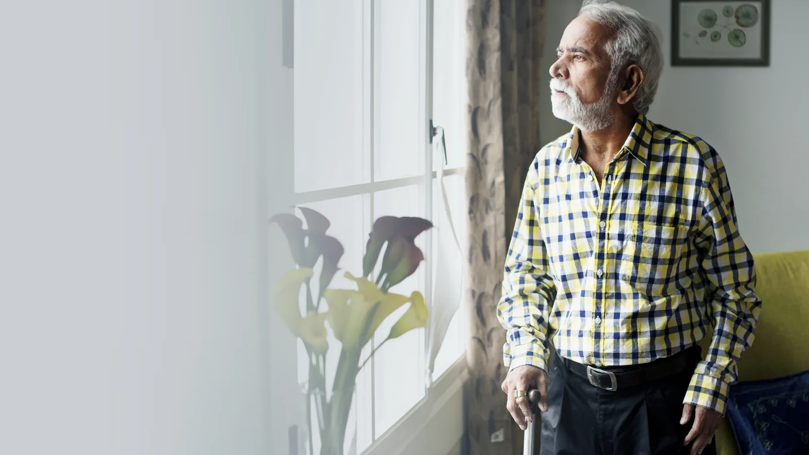 An older man standing beside a window at home, with one hand on a walking stick.