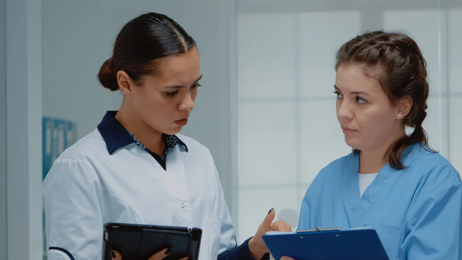 Two healthcare staff in uniform talk seriously; one holds a tablet and the other a clipboard.